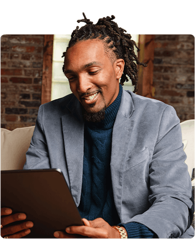 A man with grey sports coat and blue sweater is sitting down looking at his iPad