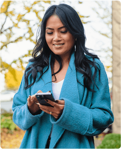 woman with dark skin and hair is outside wearing a teal coat and using her phone for mobile banking