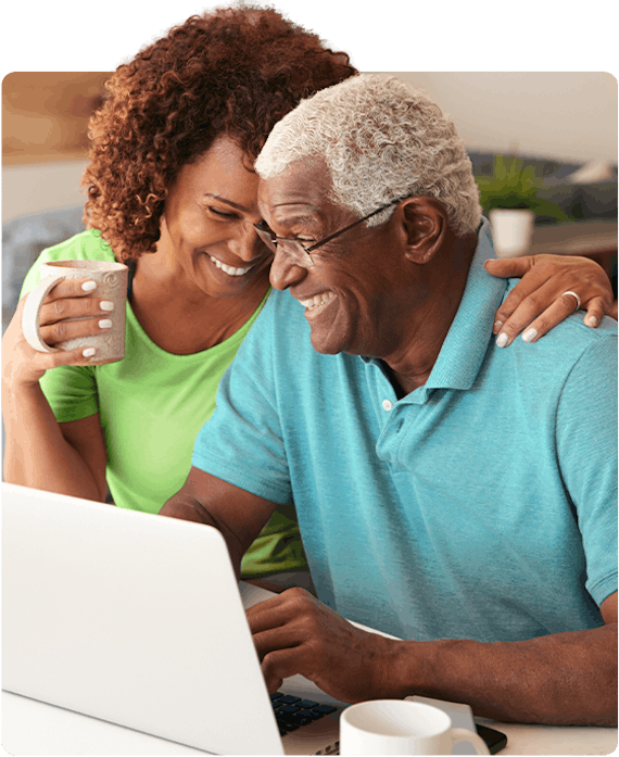 Woman wearing a light green shirt holding a cup of coffee is smiling and hugging her husband who is wearing light blue shirt while looking at laptop