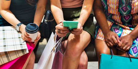 Three seated women shown from the neck down holding coffee, colorful shopping bags, and a cell phone