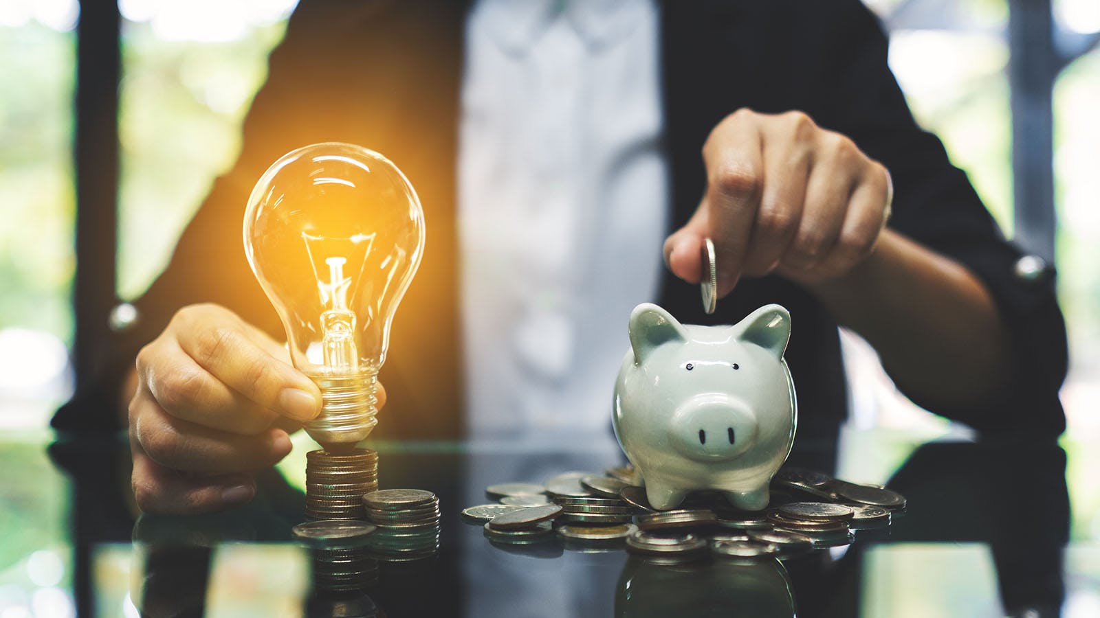 Closeup of man in suit sitting behind desk holding glowing lightbulb and filling piggy bank with coins