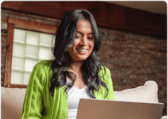 A young woman with olive skin and dark hair wearing a lime green colored sweater is sitting on a couch looking at a laptop on her lap