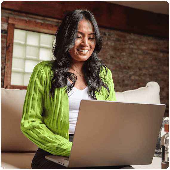 A young woman with olive skin and dark hair wearing a lime green colored sweater is sitting on a couch looking at a laptop on her lap