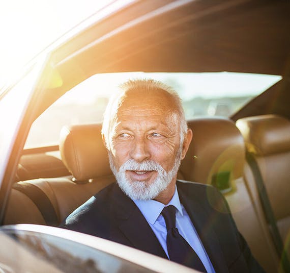 An older man wearing a nice suit is sitting in the backseat of a car looking out a rolled down window
