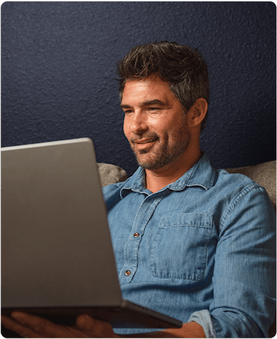 a man with dark hair is wearing a denim button up shirt and his sitting on his cough looking at his laptop