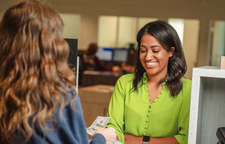 Woman with dark hair wearing a lime green blouse is at an OMB teller line receiving money