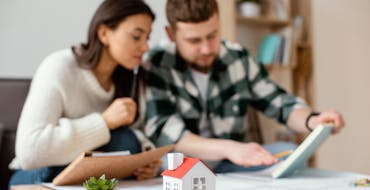 A couple flipping through paperwork with a house figurine on the desk and a small potted succulent