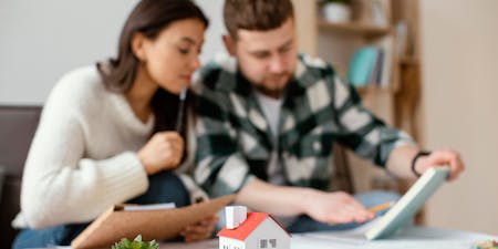 A couple flipping through paperwork with a house figurine on the desk and a small potted succulent