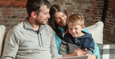 Smiling couple holding their young son on their lap while looking at a laptop screen against a brick wall background