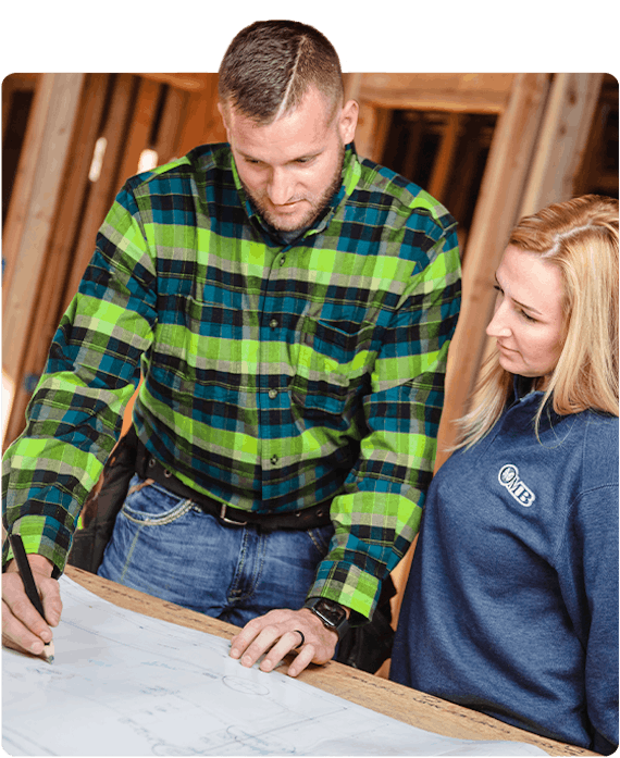 Man wearing a green and blue plaid shirt is using a carpenter pencil to make notes on a blueprint while a woman wearing blue sweater is watching intently